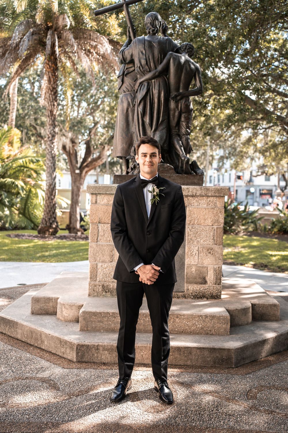 A classic black tuxedo worn in front of a bronze statue on a stone pedestal in a park setting with palm trees.