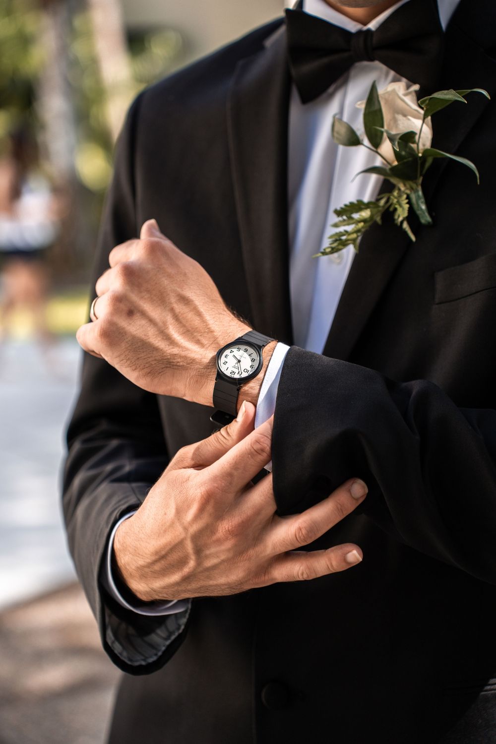 Close-up of groom in black tuxedo adjusting sleeve cuff while wearing elegant watch and white floral boutonniere.