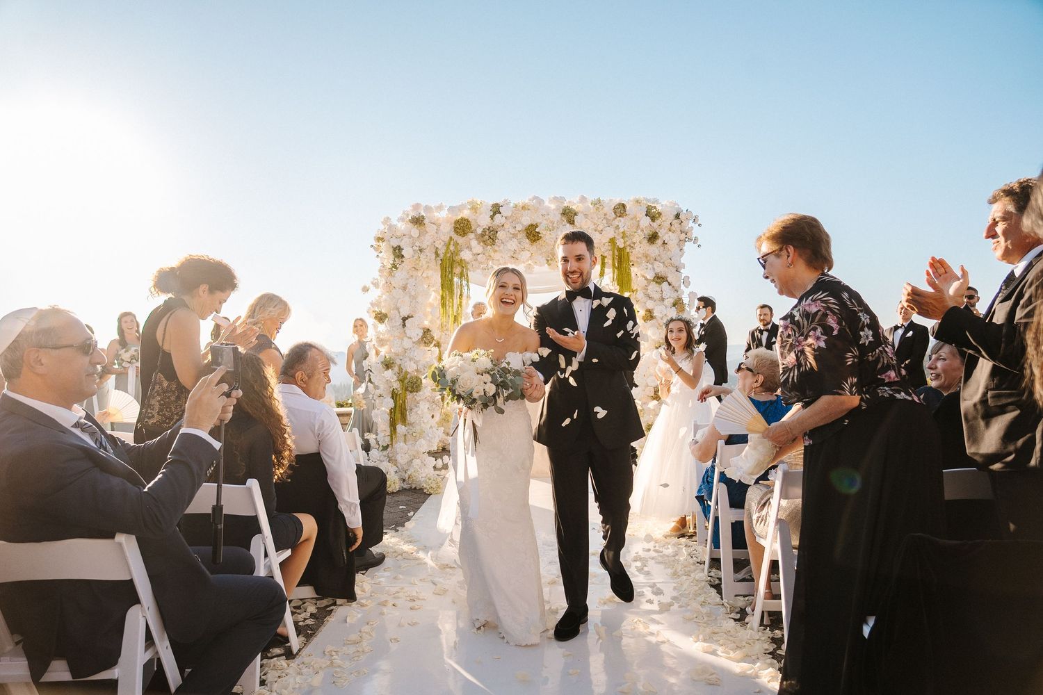 Wedding guests celebrate during outdoor ceremony with decorative floral arch and ocean backdrop at sunset.