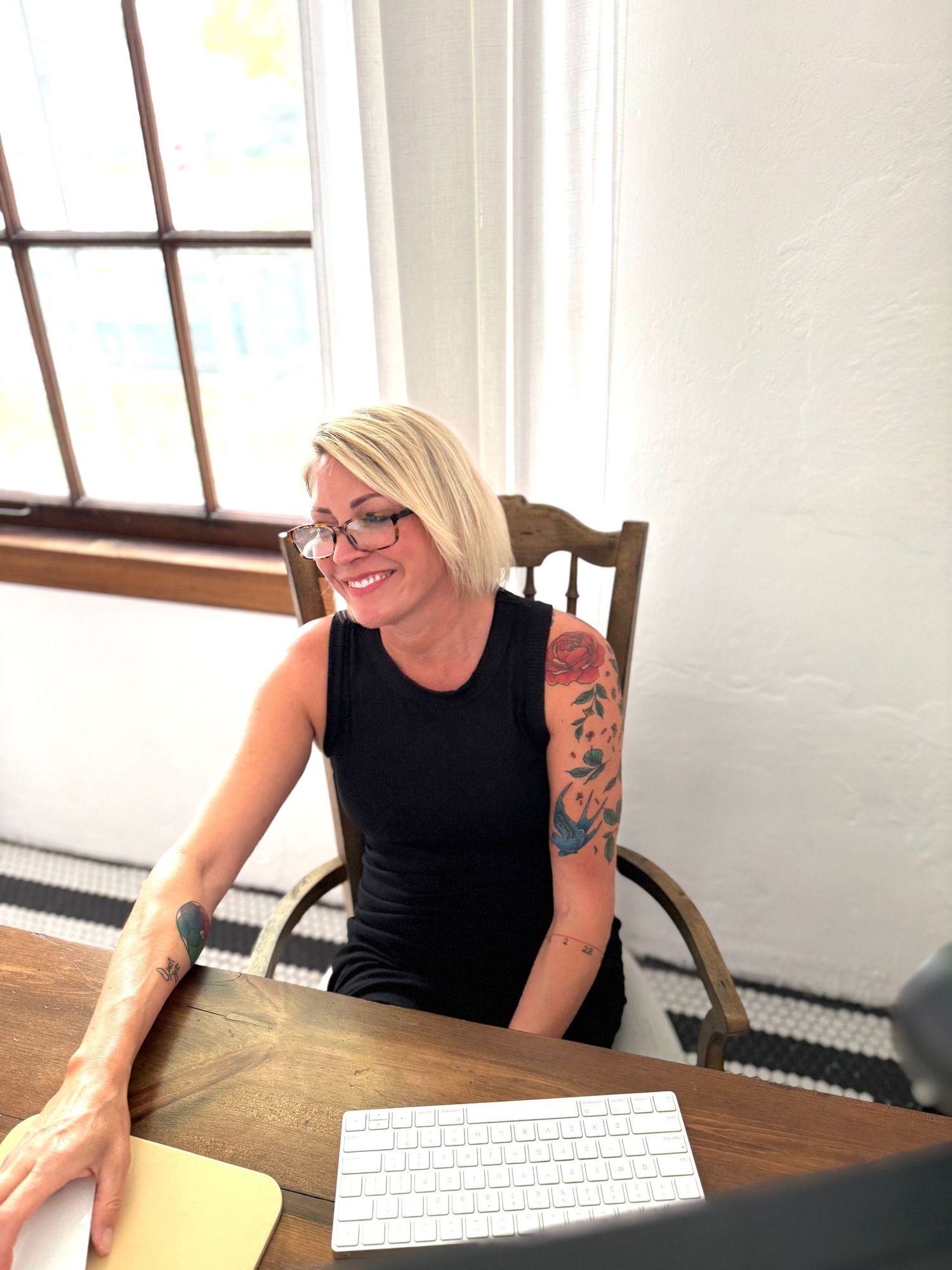 A woman with blonde hair and tattoos sits at a desk, working on a computer in a bright room.