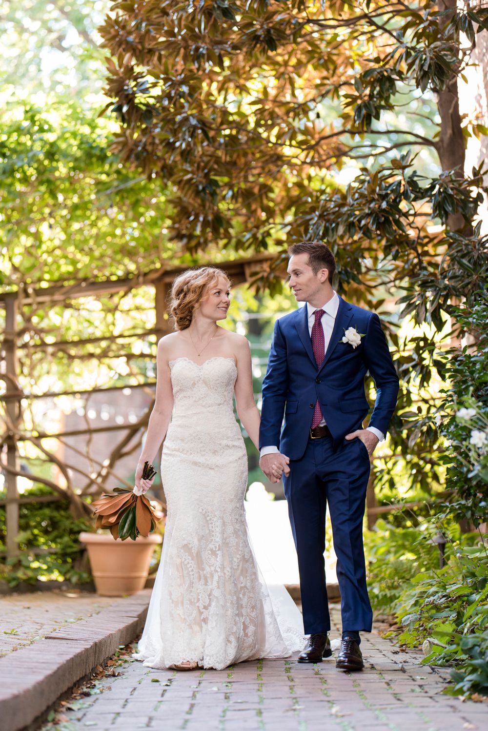 A couple walks hand in hand along a garden path, with the bride in lace and groom in navy suit under magnolia trees.