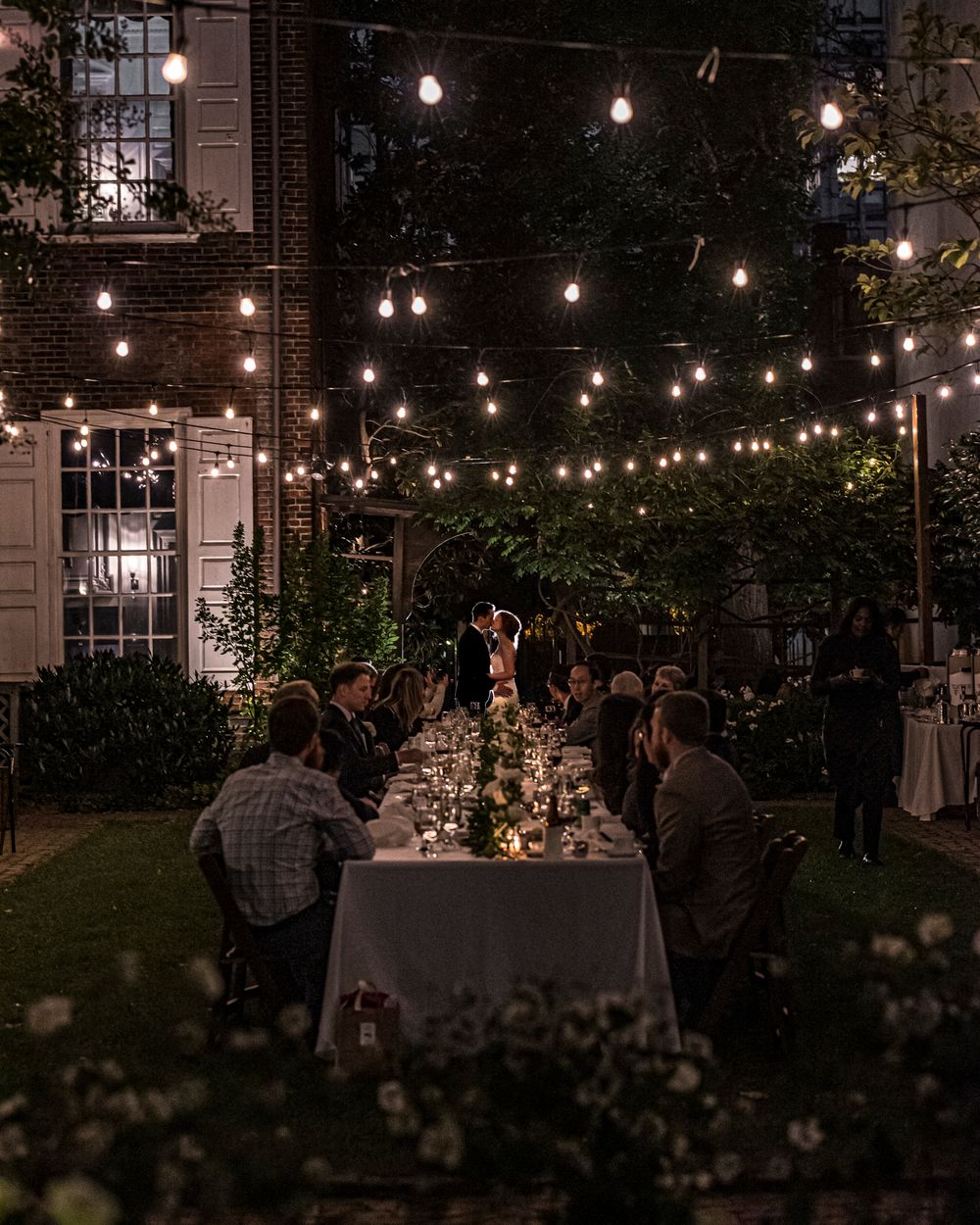 An elegant outdoor dinner party under string lights with a long white table set up in a garden at dusk.