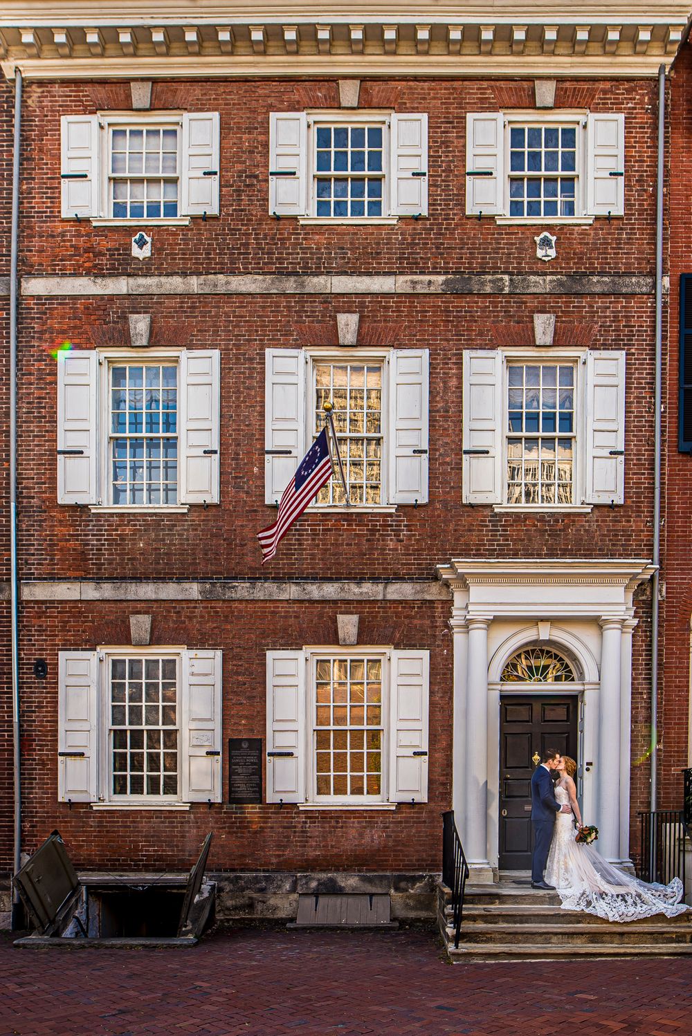 Historic brick building with white shuttered windows, American flag, and couple standing at arched doorway in Philadelphia.