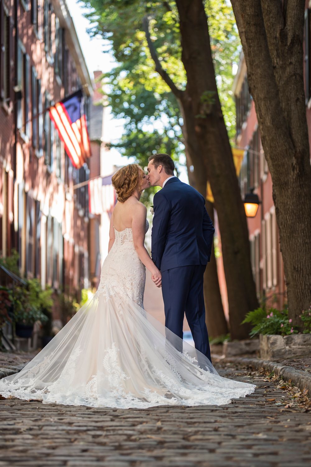 Newlywed couple in formal attire photographed on historic cobblestone street with brick buildings and trees in background.