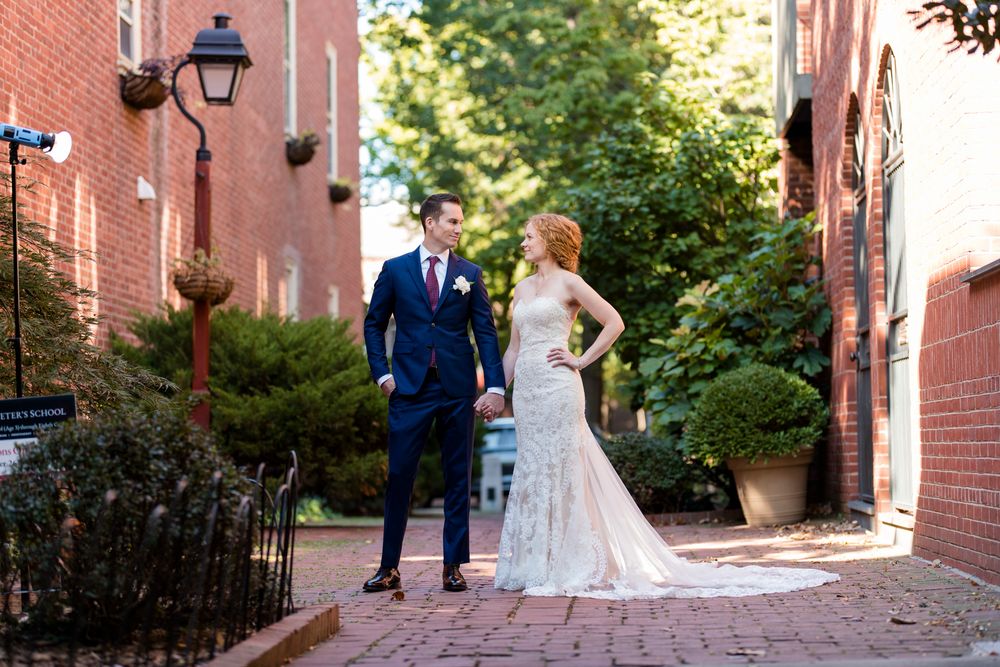 A couple in wedding attire stands on a charming brick pathway between historic buildings on their special day.