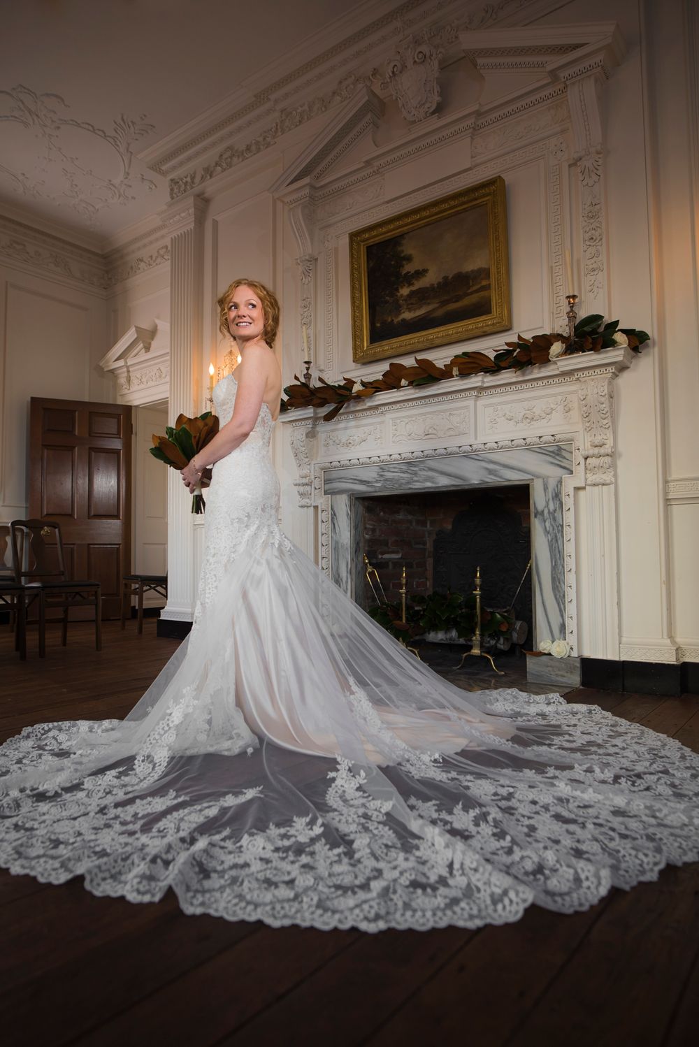 A stunning cathedral-length lace wedding dress train spreads elegantly across a grand room with ornate white walls and fireplace.