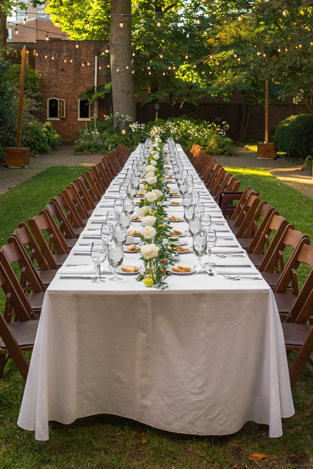 Long white dining tables elegantly set with place settings and chairs arranged outdoors under green trees.