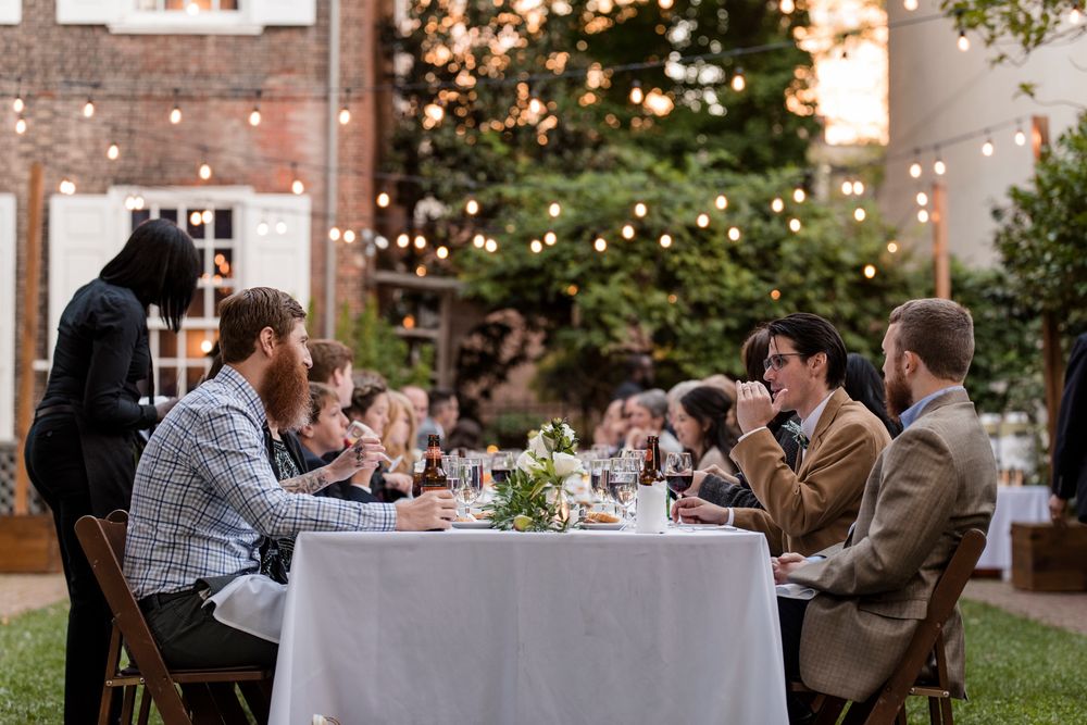 Wedding guests enjoy a candlelit outdoor reception at dusk with string lights illuminating tables and brick walls.