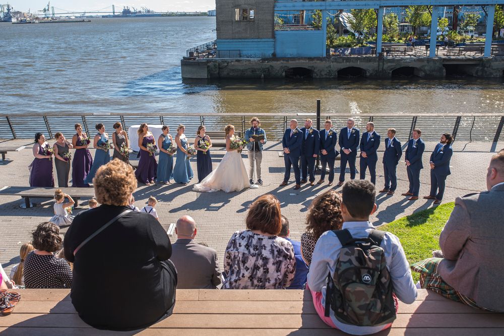 A group of people gathered along a waterfront viewing area with boats and harbor scenery in the background.