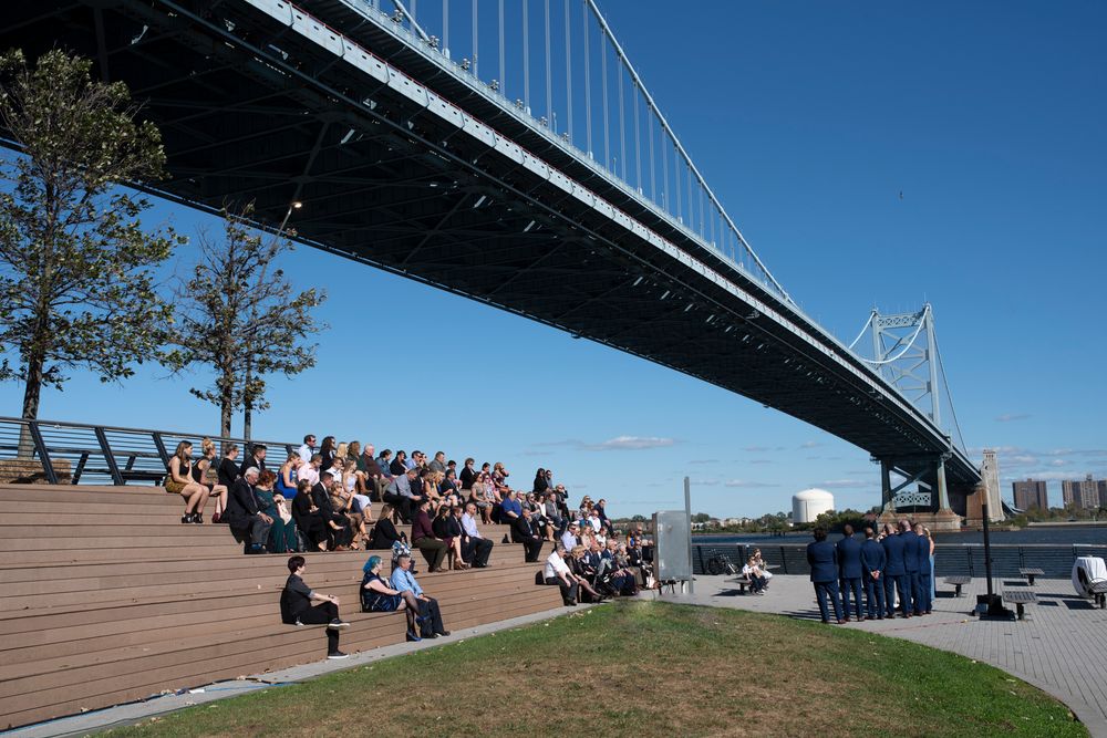 People gather on benches beneath a suspension bridge on a sunny day along the waterfront.