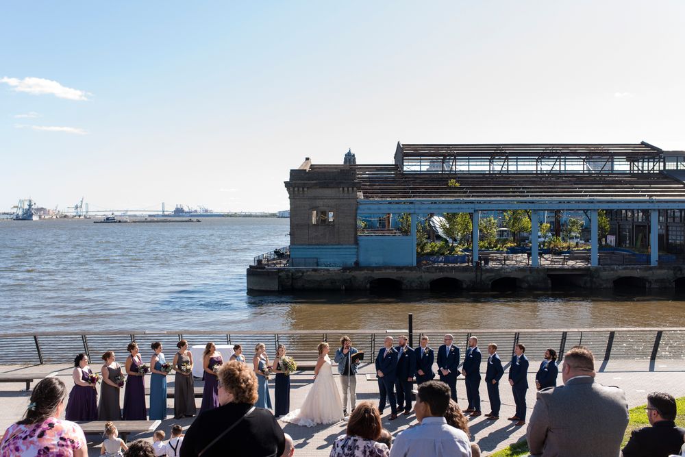 A panoramic view of people standing along a pier by the ocean with buildings and boats visible in the distance.