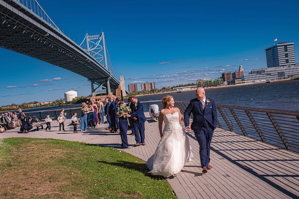A couple poses for wedding photos along the waterfront with the Benjamin Franklin Bridge in Philadelphia visible.