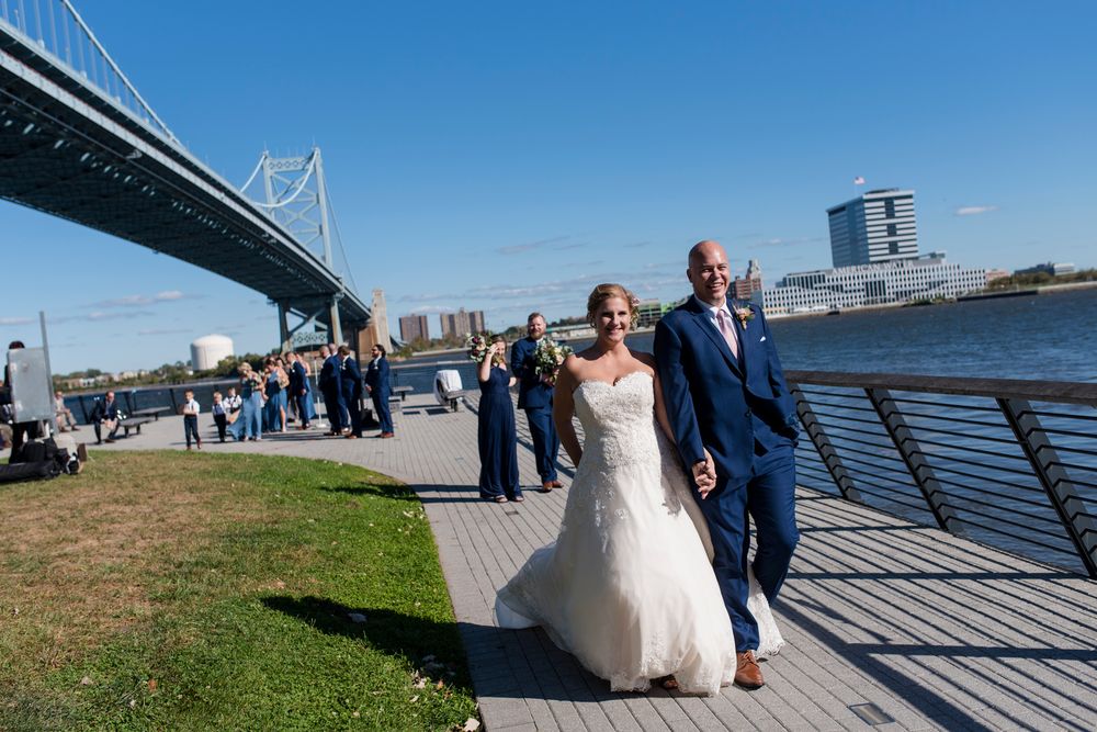 Wedding party poses along waterfront boardwalk with city skyline and bridge in background on sunny day.
