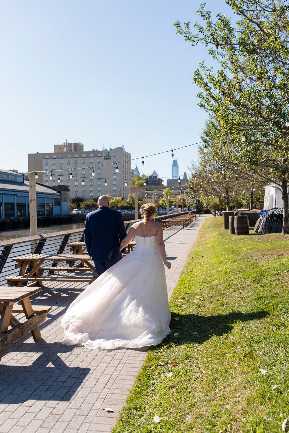 A couple in wedding attire walk along a paved pathway with picnic tables and city buildings in the background.