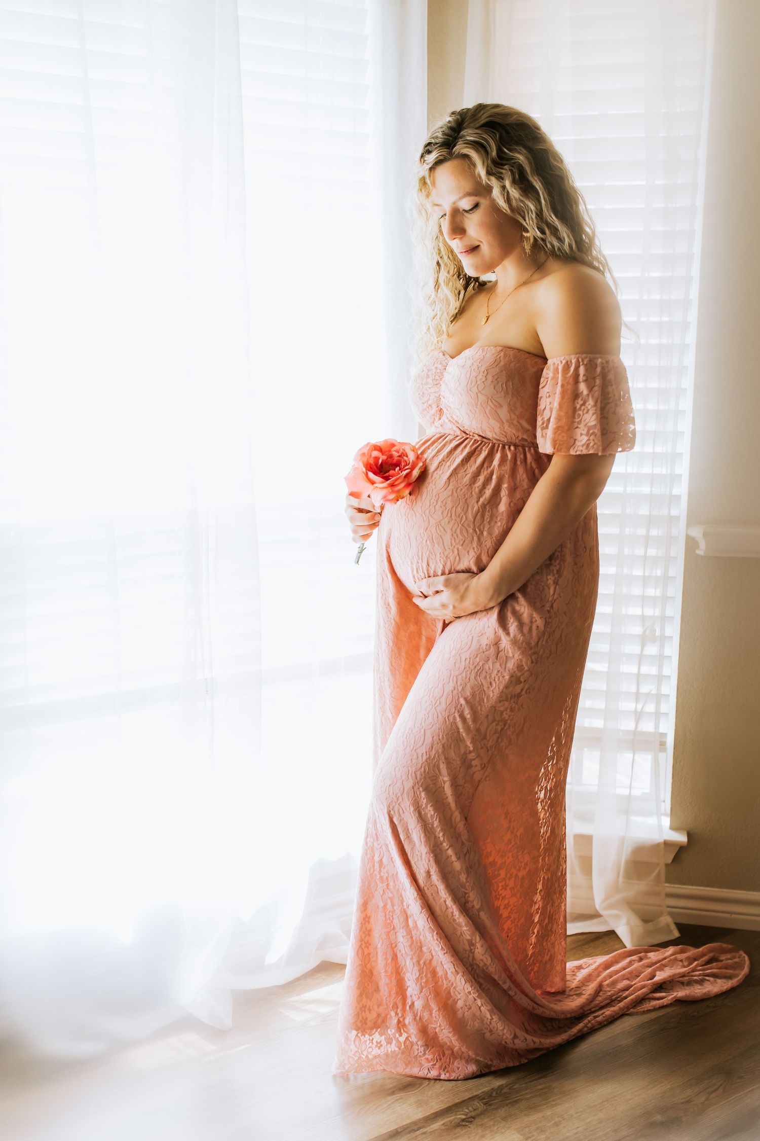 Maternity photo showing a pregnant woman in a pink lace dress posing gracefully by a sunlit window in Keller, TX.
