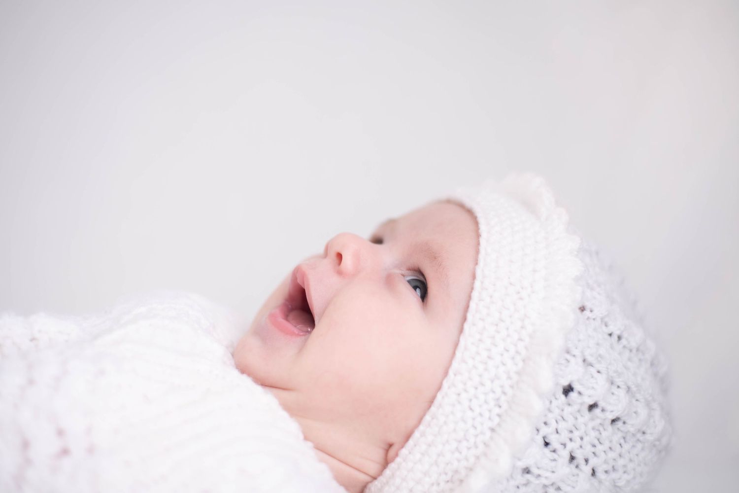Adorable baby wearing a white knit hat looking upward with mouth open against a light background.