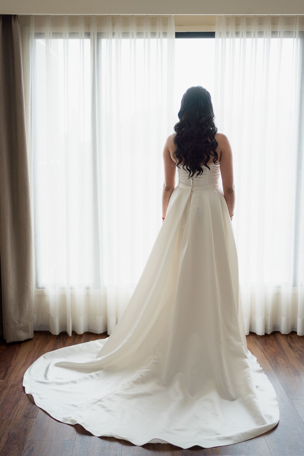 Back view of a bride in a flowing white wedding gown with a long train standing against sheer curtains in natural light.
