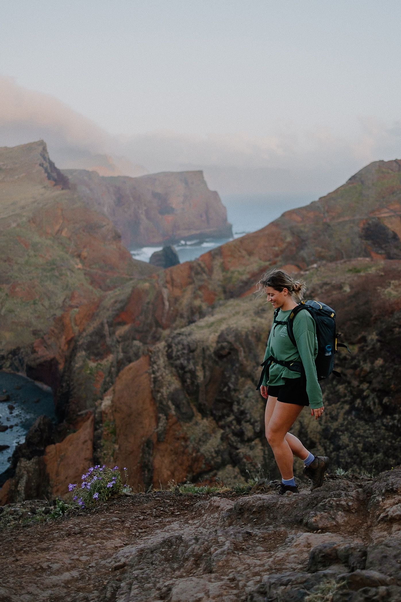 Hiker with backpack traverses rugged coastal cliffs at sunset along dramatic mountain ridges overlooking the ocean.