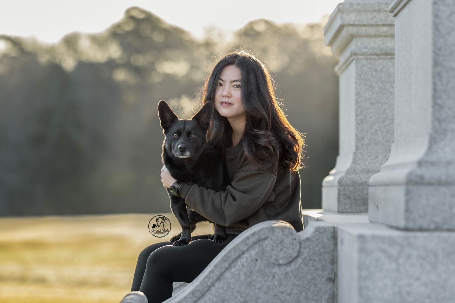 A black dog and person sit together outdoors at sunset, creating a warm silhouette against the golden light.