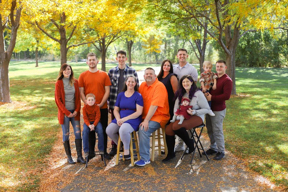 A group poses for a fall family photo outdoors with golden autumn leaves and trees in the background.