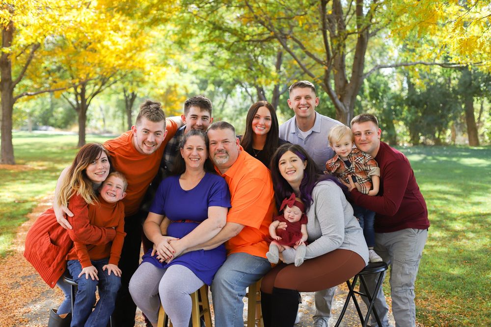 Group of people in autumn colors pose together for outdoor family photos in a park setting with yellow leaves.