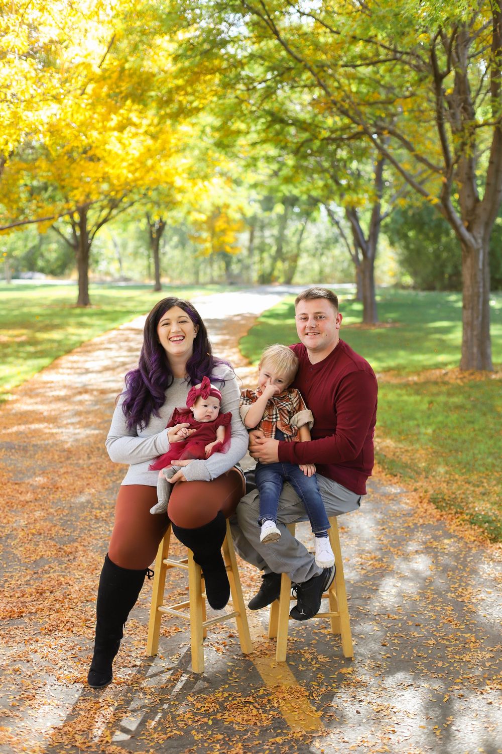 Family sits on wooden stools along autumn path with golden leaves and trees in park setting.