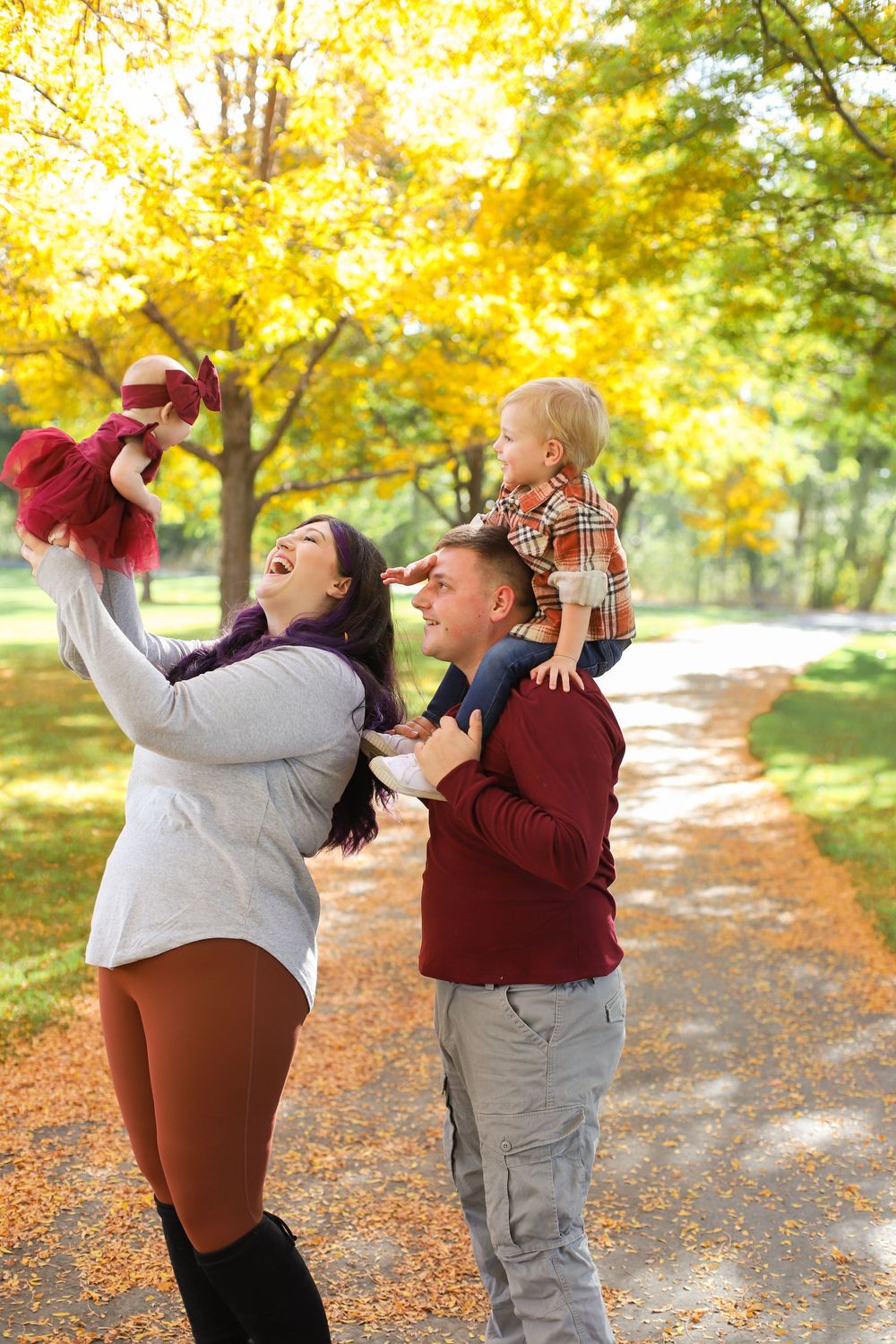 Two people playfully toss autumn leaves in the air while walking along a tree-lined path on a sunny fall day.