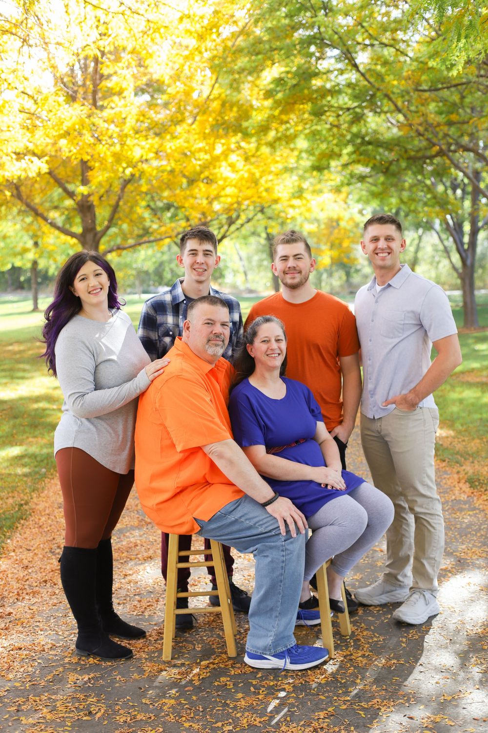 A family group poses together on a bench surrounded by golden autumn leaves in an outdoor setting.