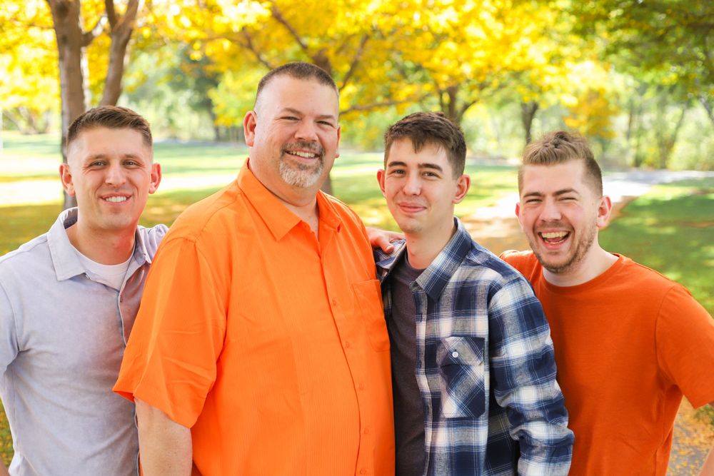 A group in casual attire smiles together outdoors with autumn foliage and trees in the background.