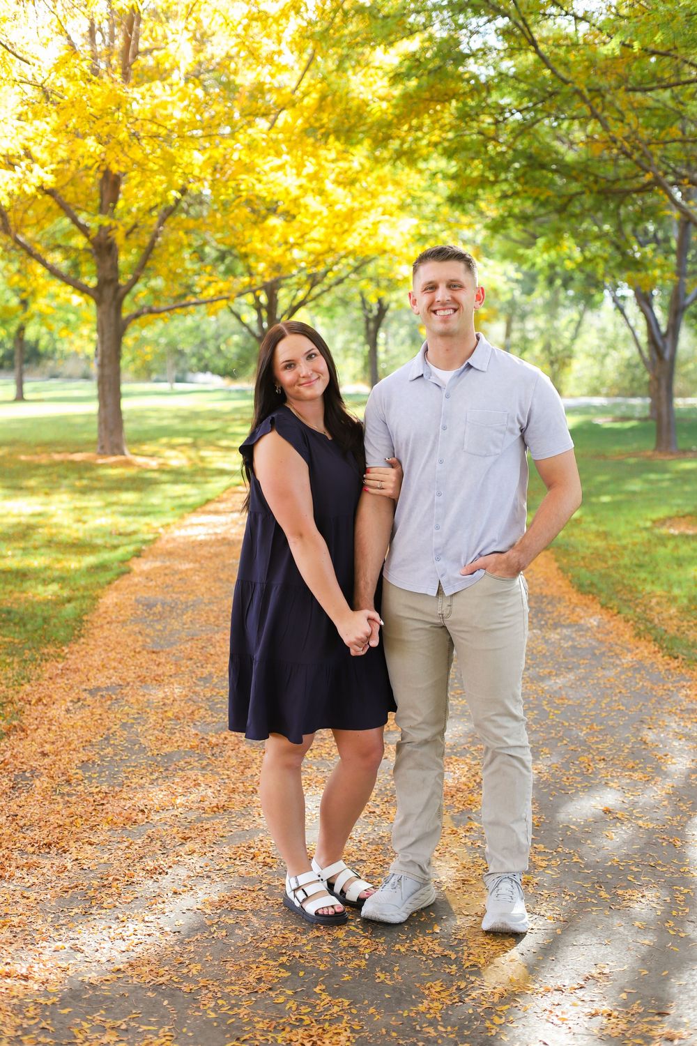 A couple poses together on a tree-lined path covered in golden autumn leaves on a sunny fall day.