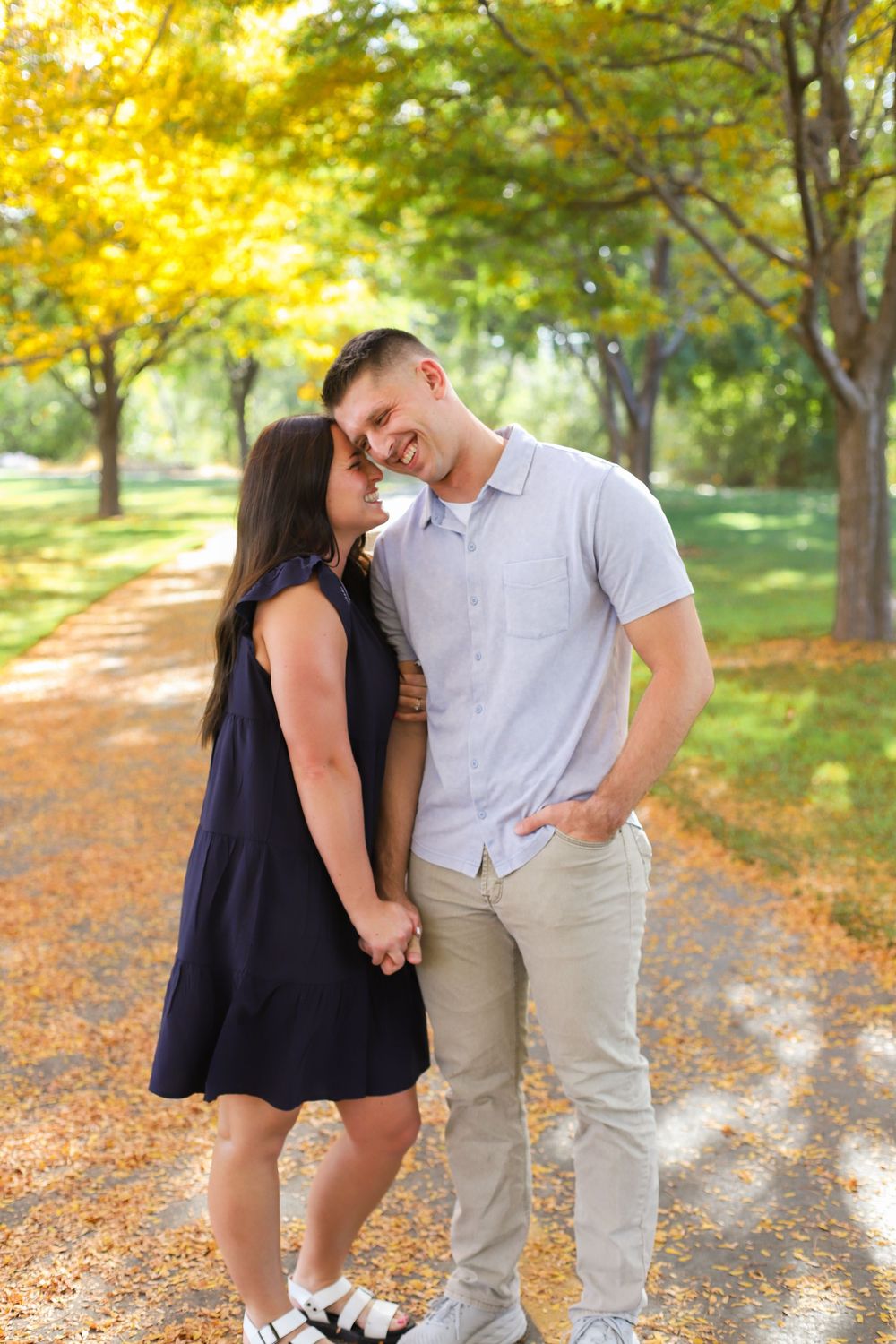 A couple shares intimate moments in a sunlit autumn park with golden leaves and trees in the background.