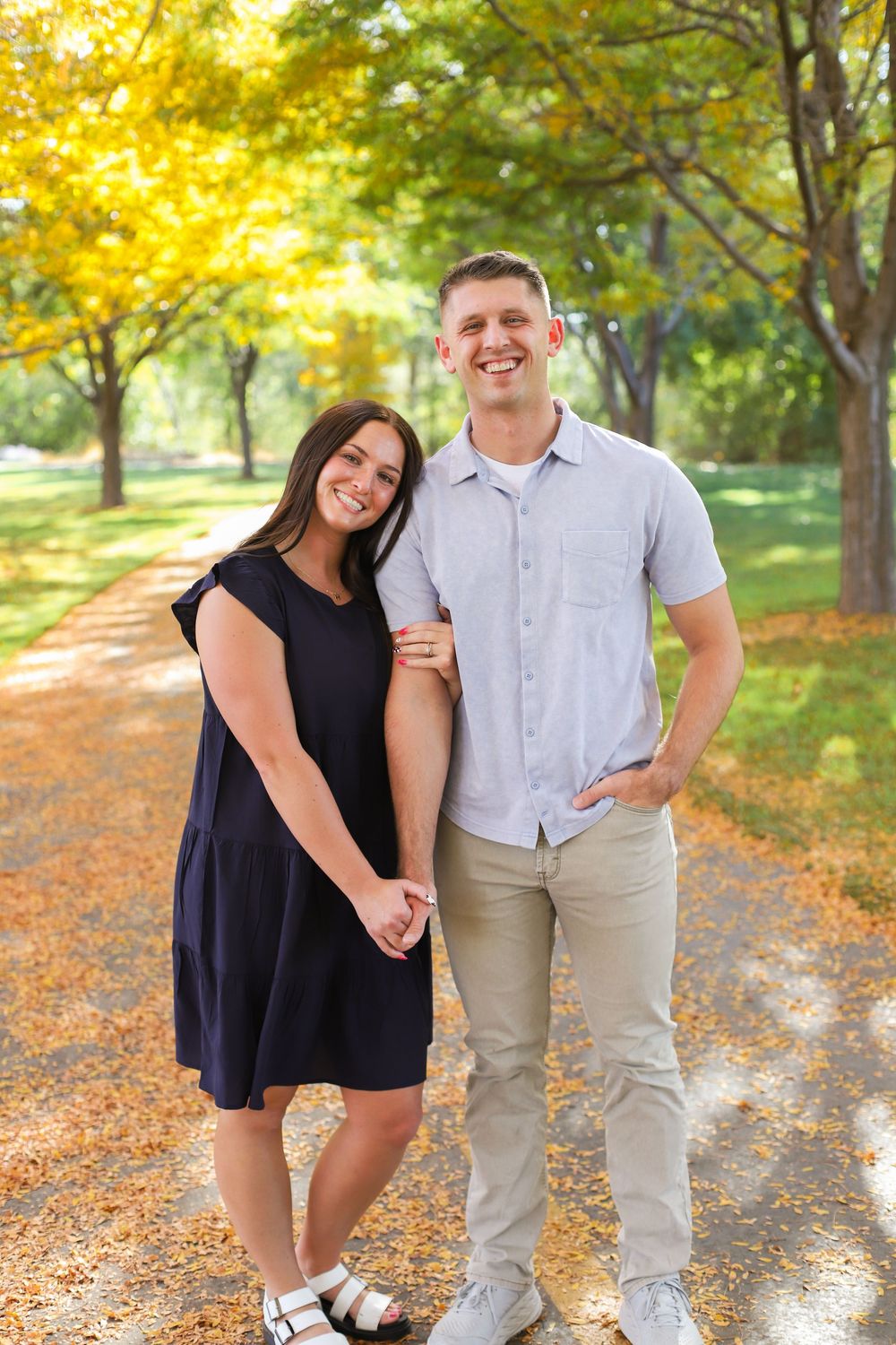 Couple enjoying a playful autumn photoshoot among golden trees and fallen leaves in a sunlit park.