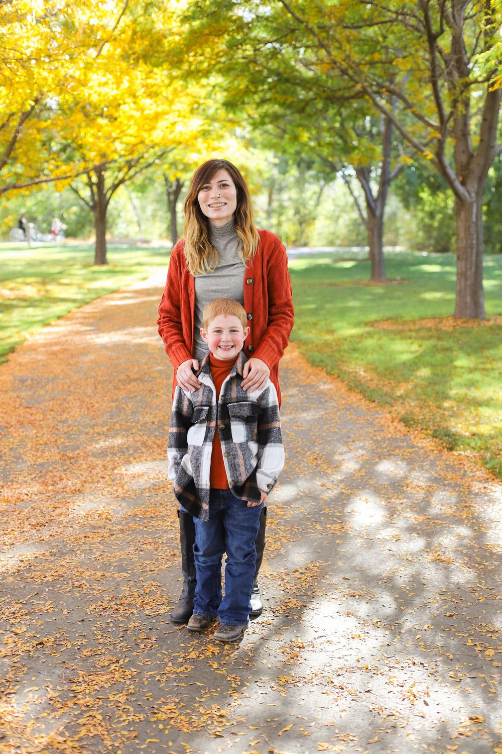 A parent and child stand together on a tree-lined autumn path with golden foliage overhead and fallen leaves below.
