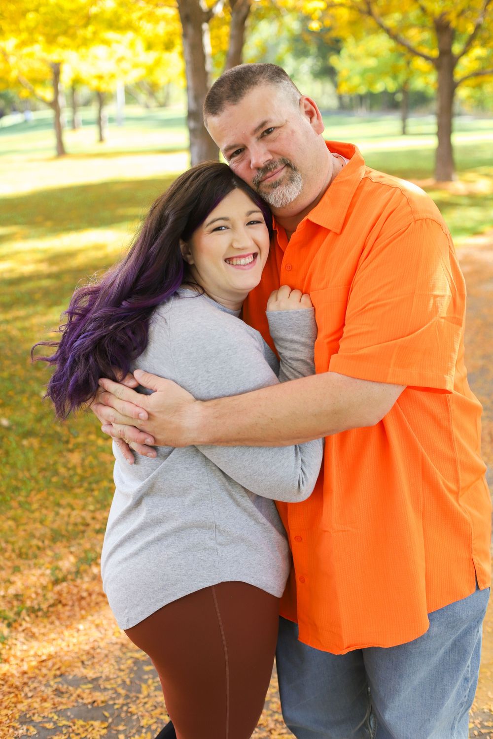 A couple embraces and laughs together during an autumn photoshoot with yellow trees and fallen leaves in the background.