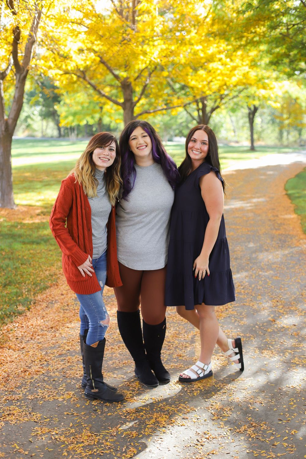 Three friends pose together on a leaf-covered path during autumn with golden yellow foliage in the background.