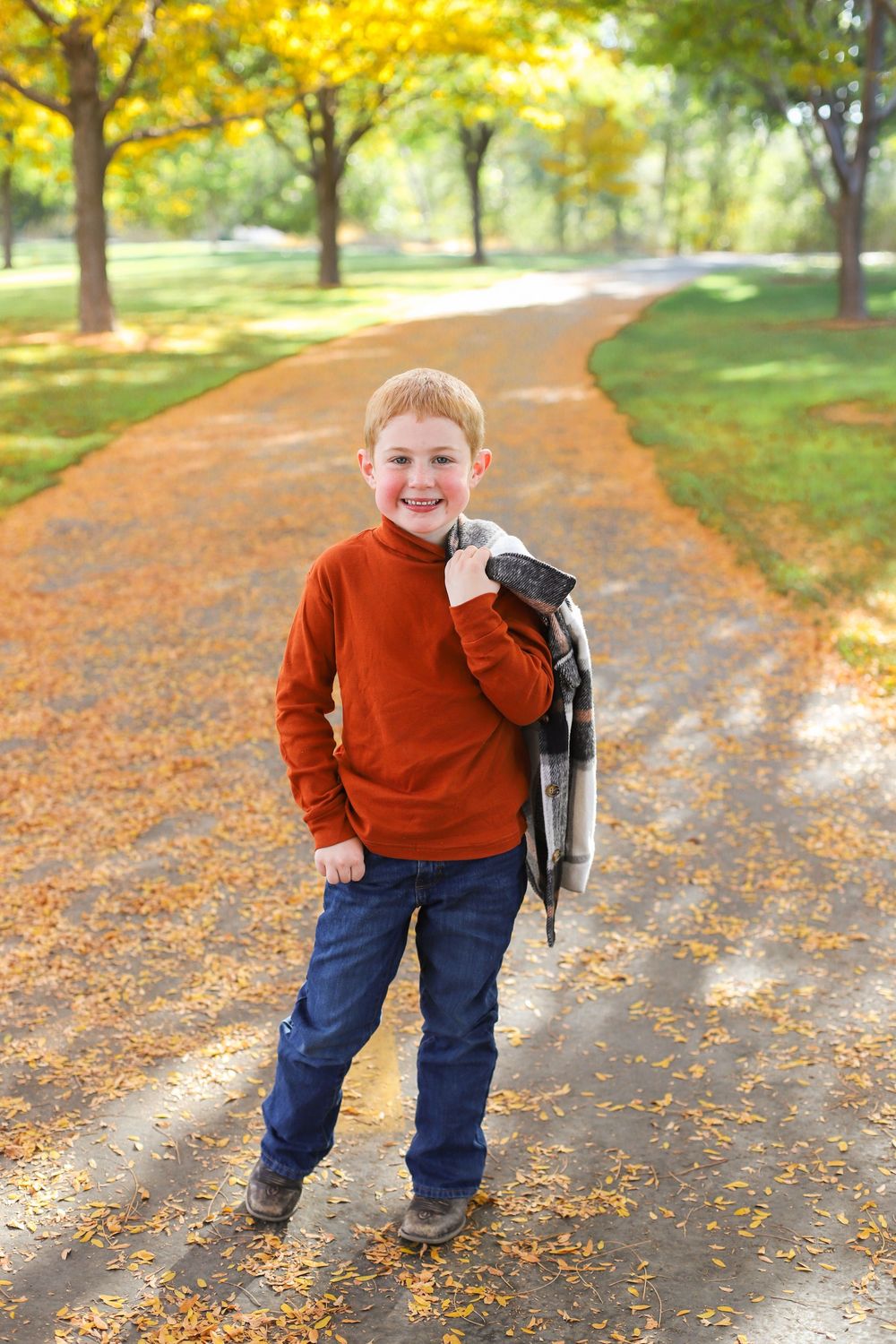 A child plays in fallen autumn leaves along a sunlit path surrounded by golden trees.