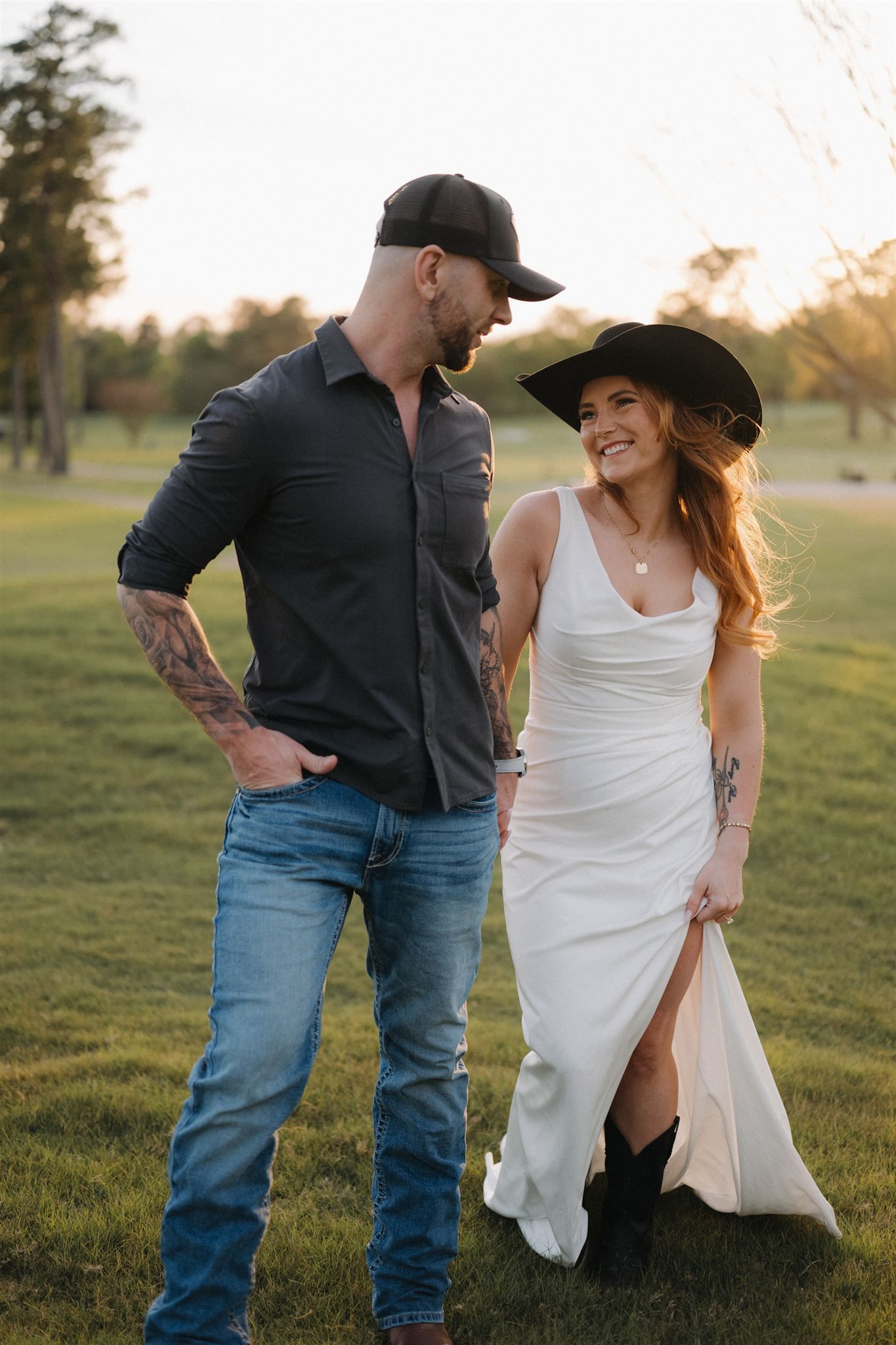A couple in casual and formal attire playfully poses together in a grassy field during golden hour.
