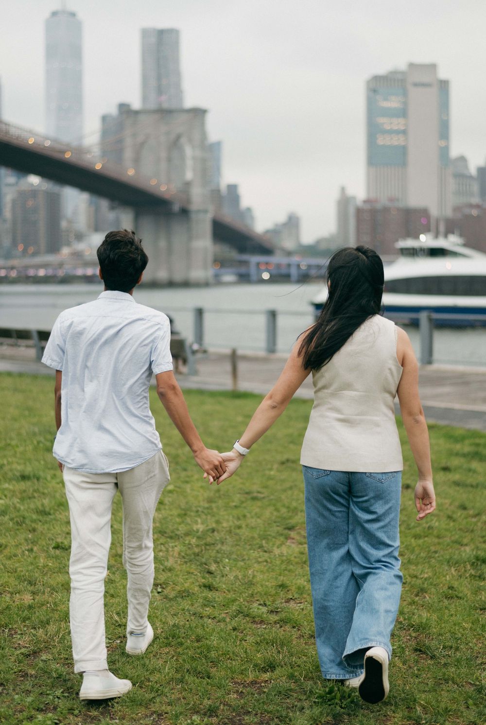 Couple walking hand in hand on grassy lawn with city skyline and waterfront in the background.