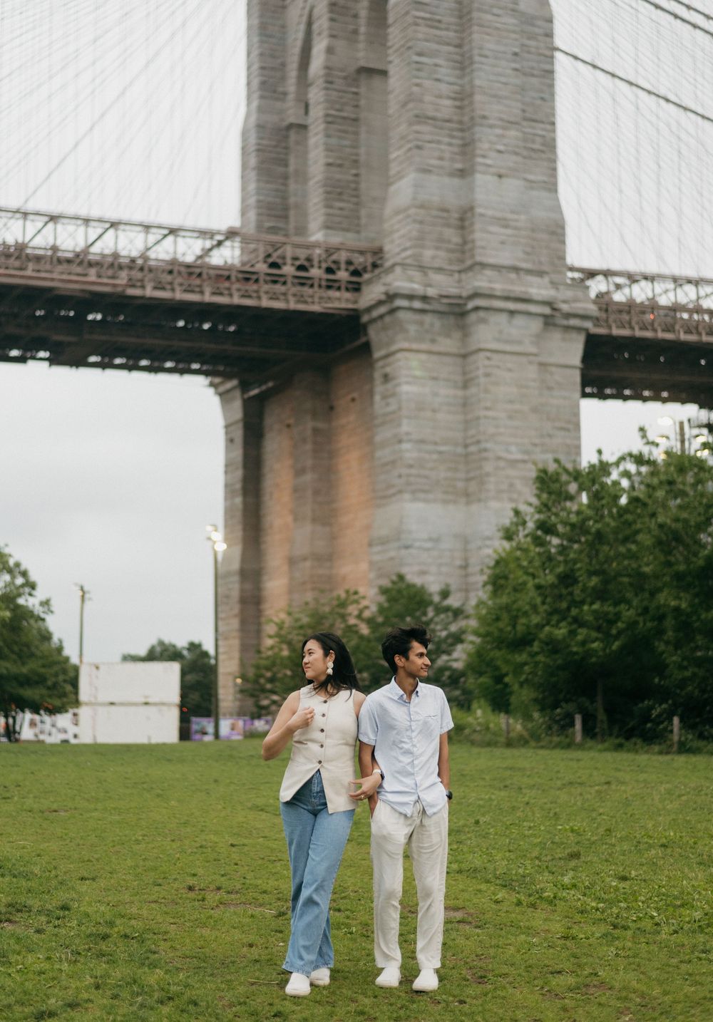 A couple walks together on a grassy hill beneath the towering stone pillars of a bridge.
