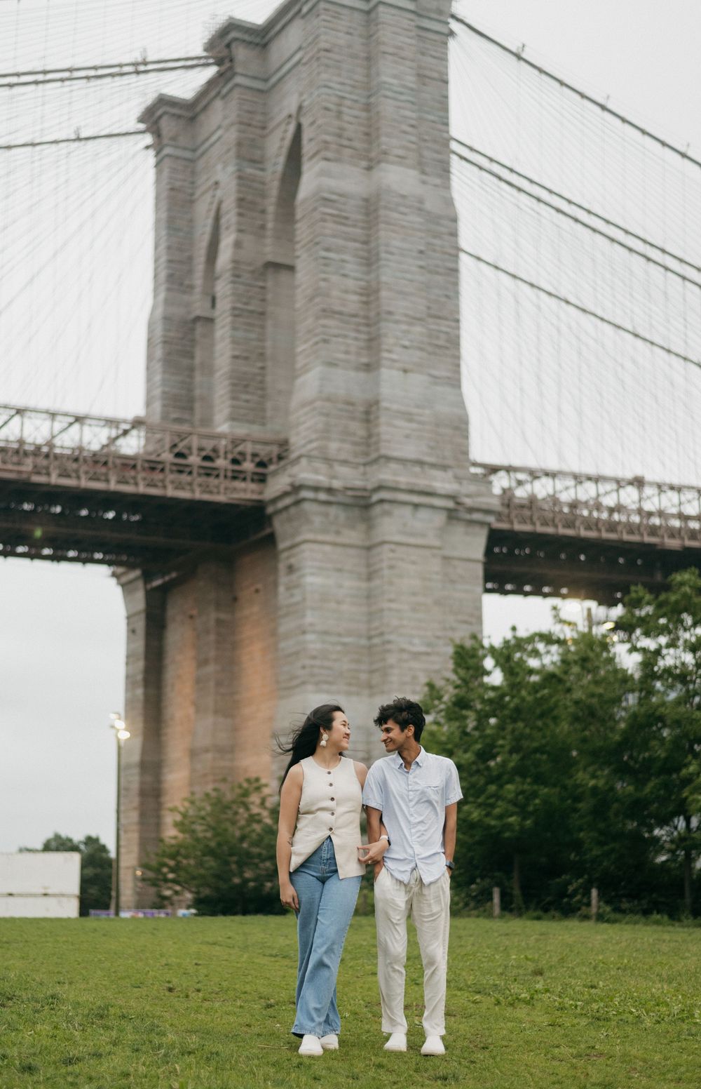 Couple standing together on a grassy hill near the Brooklyn Bridge on a cloudy day.