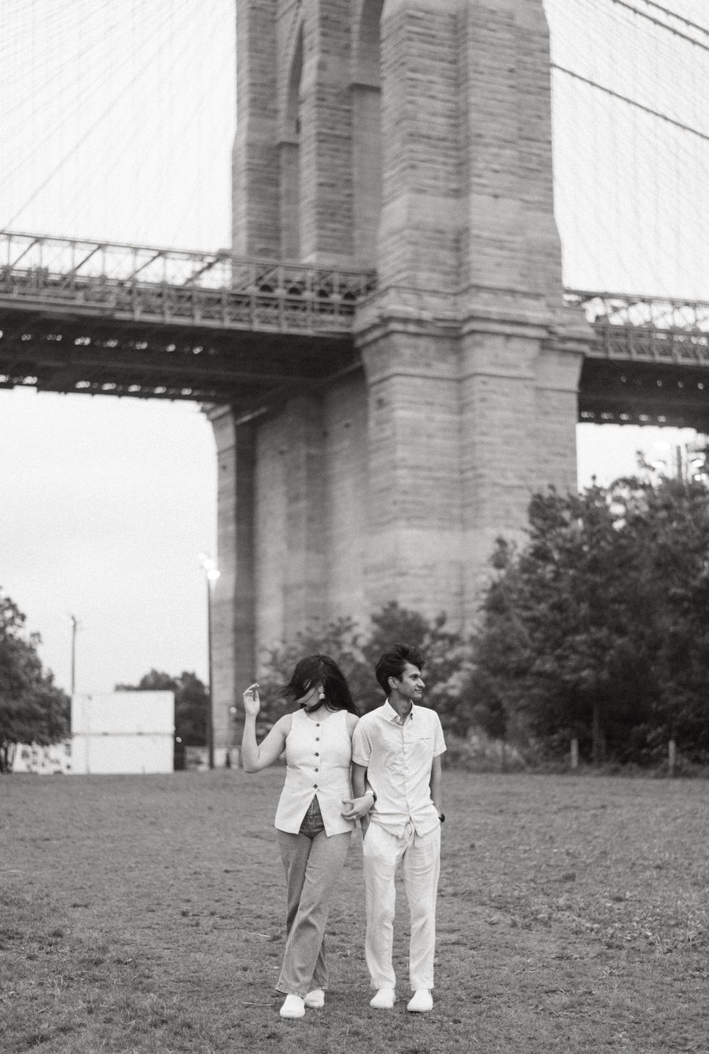 Black and white photo of a couple in white outfits standing on a grassy hill beneath a large bridge structure.