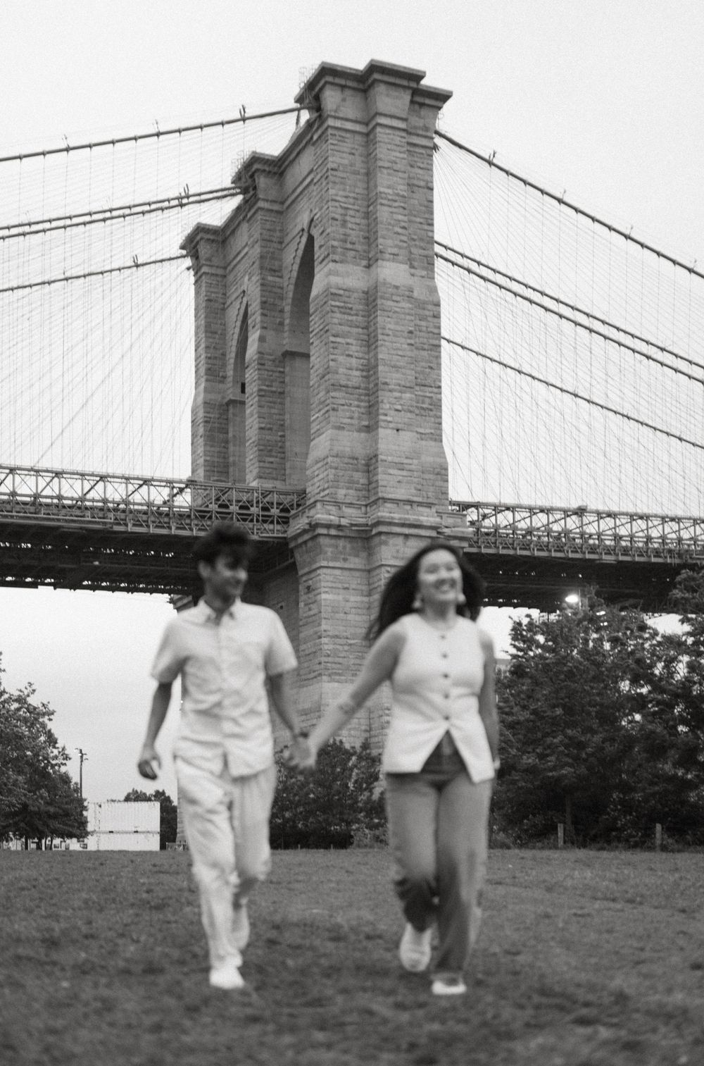 A couple in white outfits runs playfully across a grassy hill near the Brooklyn Bridge in black and white photography.