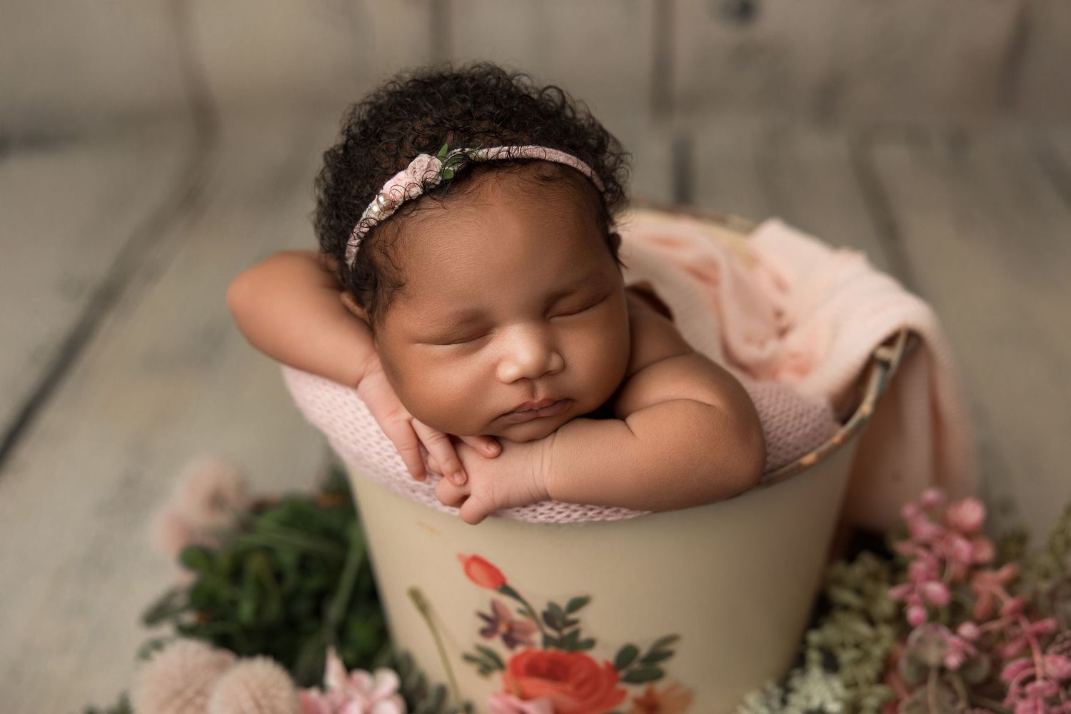 A sleeping newborn baby in a cream-colored bowl surrounded by pink flowers and greenery in a studio setting.