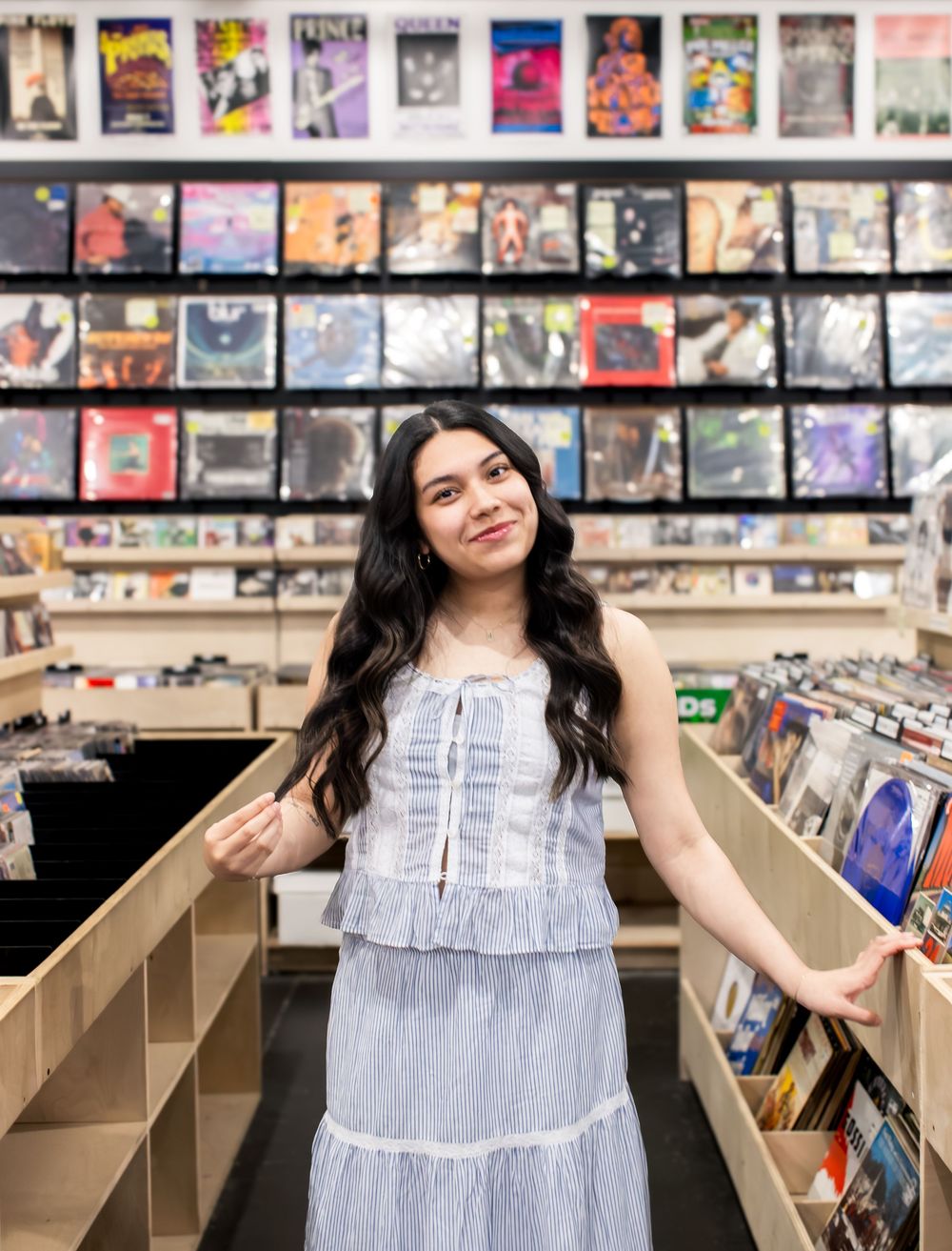 Young woman in blue dress smiling in a vinyl record store, surrounded by shelves of album covers.