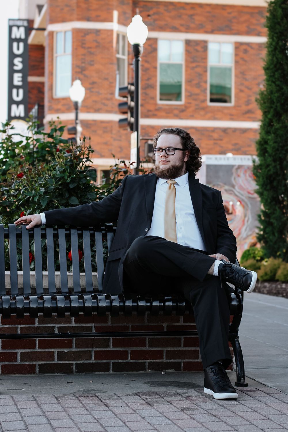 A person in a black suit and tie sits casually on a brick and metal bench outside a building.