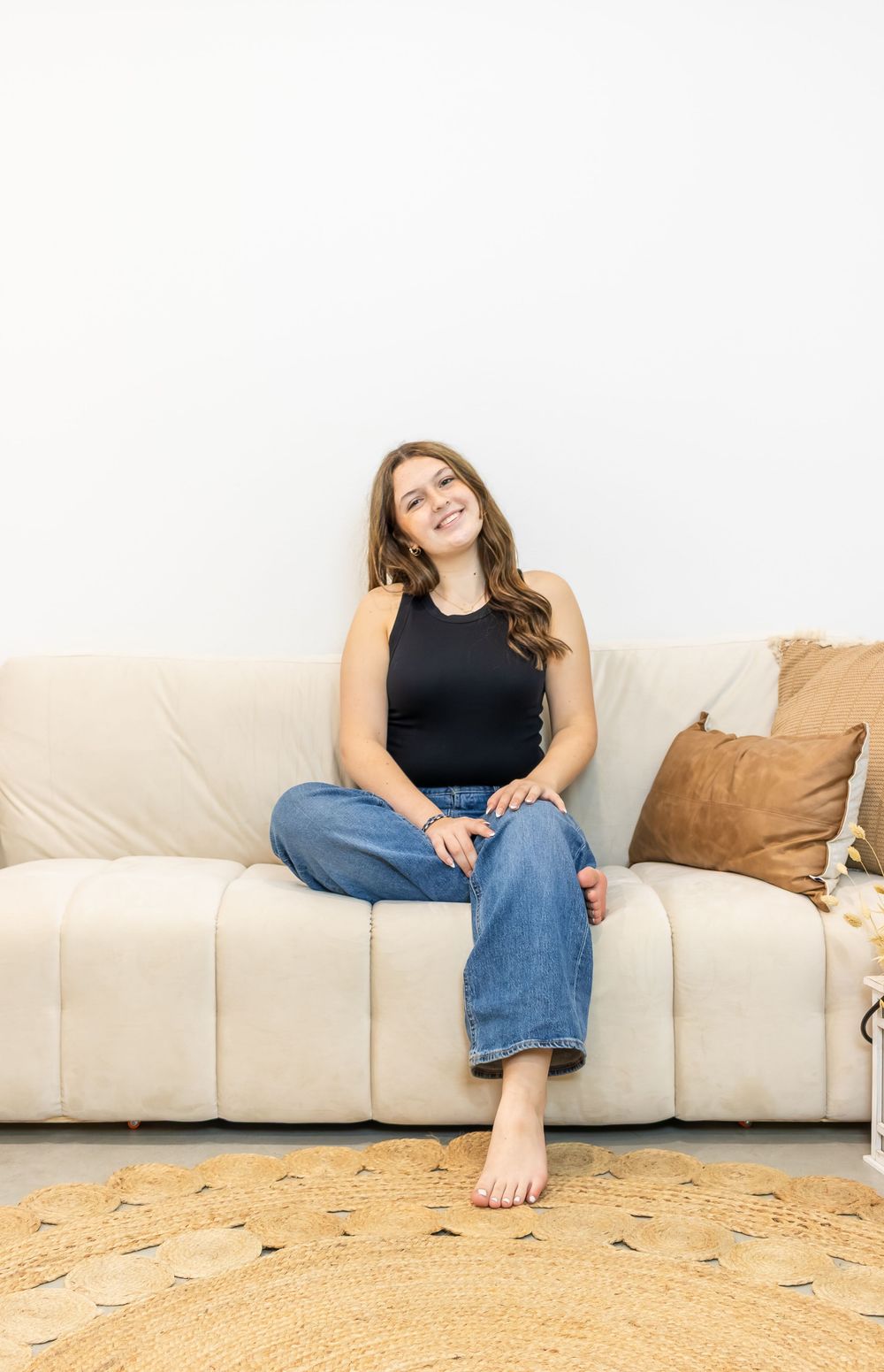 Smiling young woman in black tank top and jeans sitting cross-legged on a cream sofa with boho cushions.