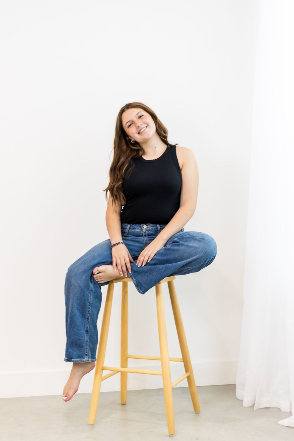 Casual lifestyle portrait of someone sitting on wooden barstool wearing black tank top and blue jeans against white wall.