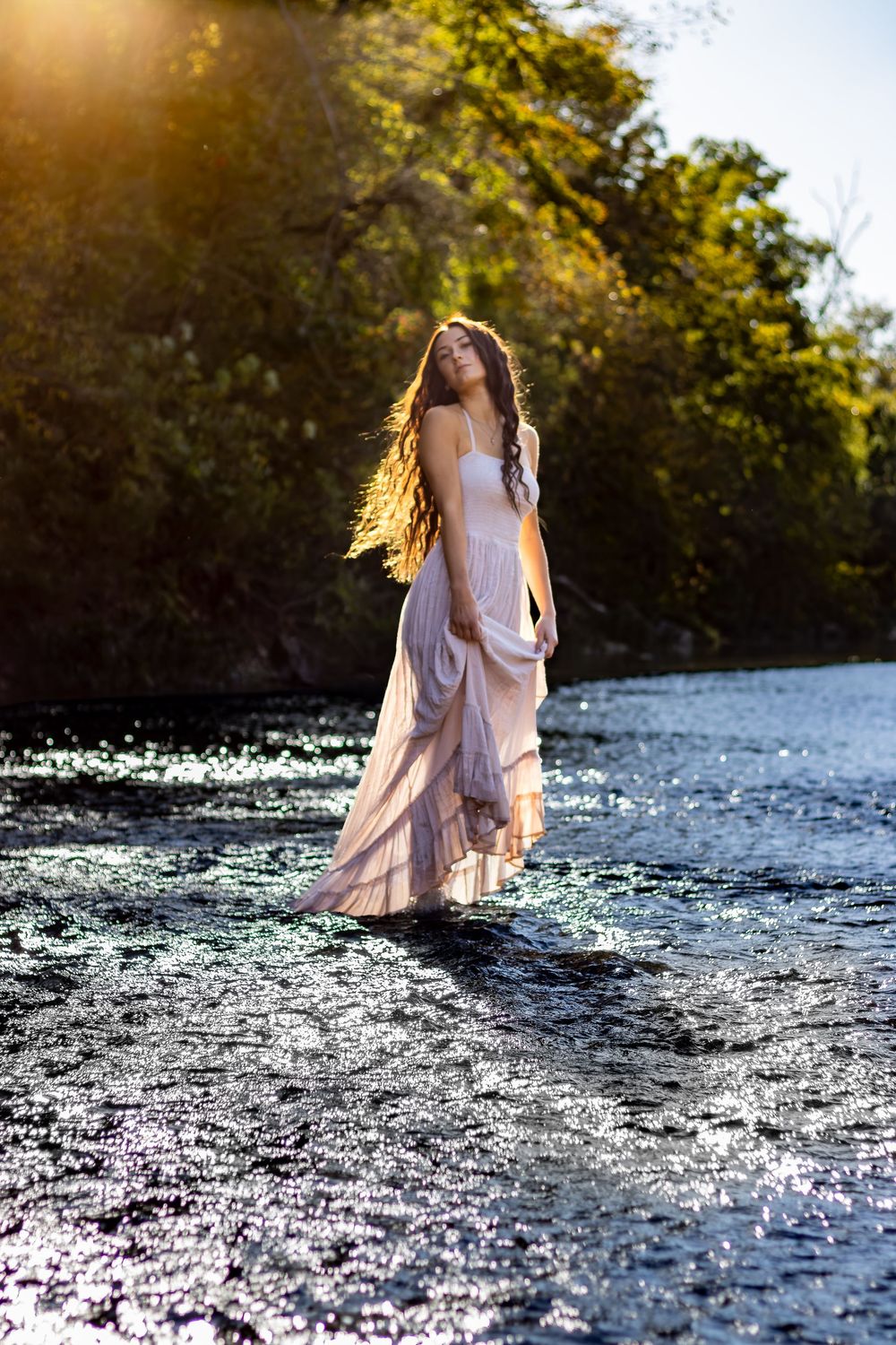 Woman in flowing pink dress standing in sunlit river with lush green trees in background.