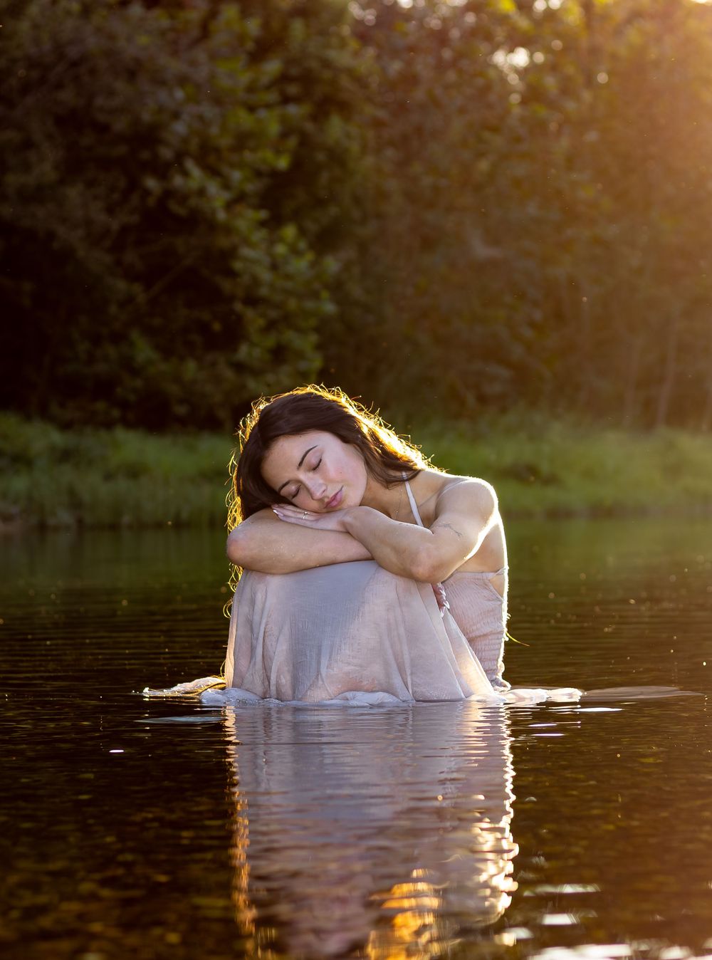 A figure in a flowing white dress poses gracefully in shallow water during golden hour sunset creating ethereal reflections.