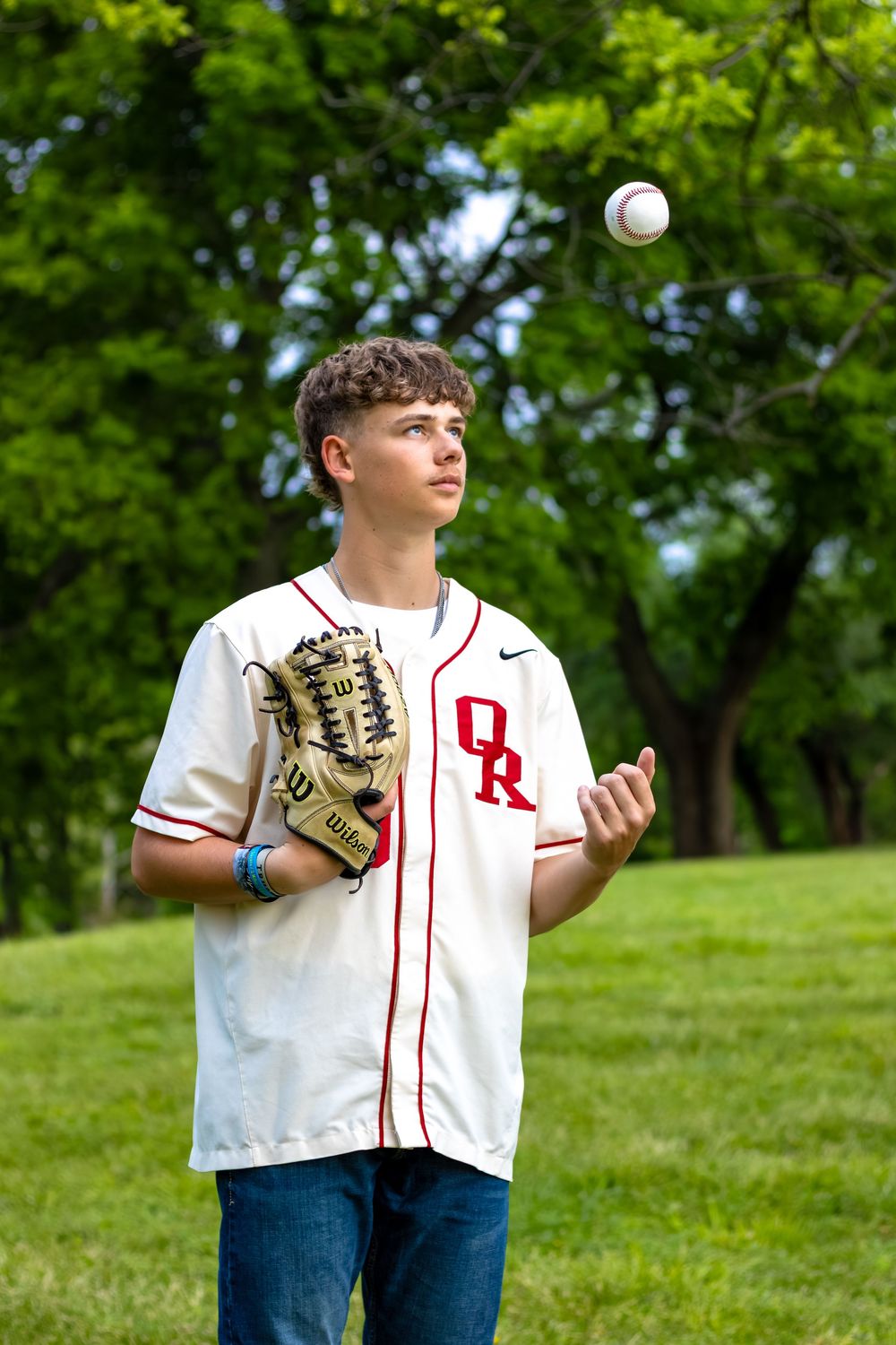 Teen baseball player in white OR jersey tossing a ball while holding a glove outdoors in a green park.